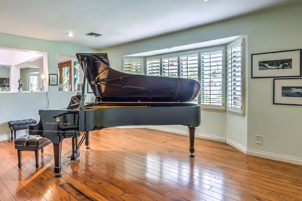 A spacious room with wooden flooring featuring a black Steinway & Sons grand piano and matching piano bench. The room has a large bay window with white plantation shutters allowing natural light to fill the space. Two framed black and white photographs hang on the wall to the right.