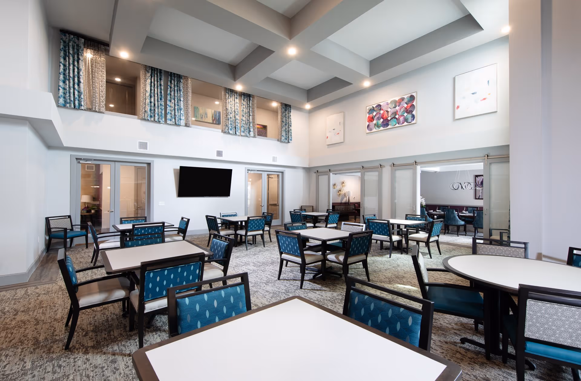 A spacious, well-lit dining room with multiple tables and chairs arranged neatly. The chairs have blue patterned upholstery. The room features high ceilings with recessed lighting, large windows with blue and white curtains, and abstract artwork on the walls. There is a flat-screen TV mounted on one wall and sliding glass doors leading to another room.