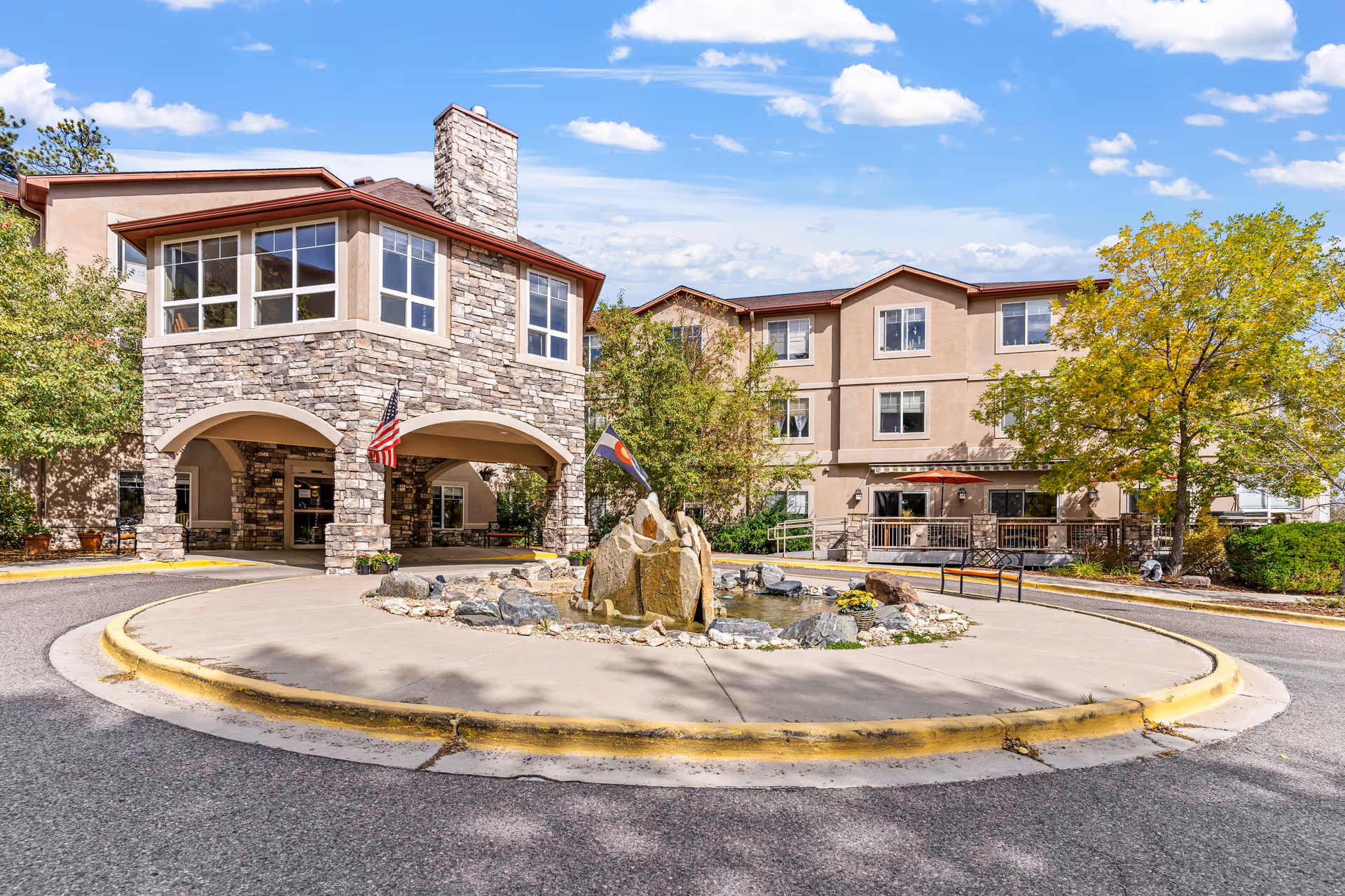 Exterior view of Elk Run Assisted Living facility showing a three-story building with stone and stucco facade, a covered entrance with arches, an American flag, a circular driveway with a rock fountain in the center, and trees with green and yellow leaves under a partly cloudy blue sky.