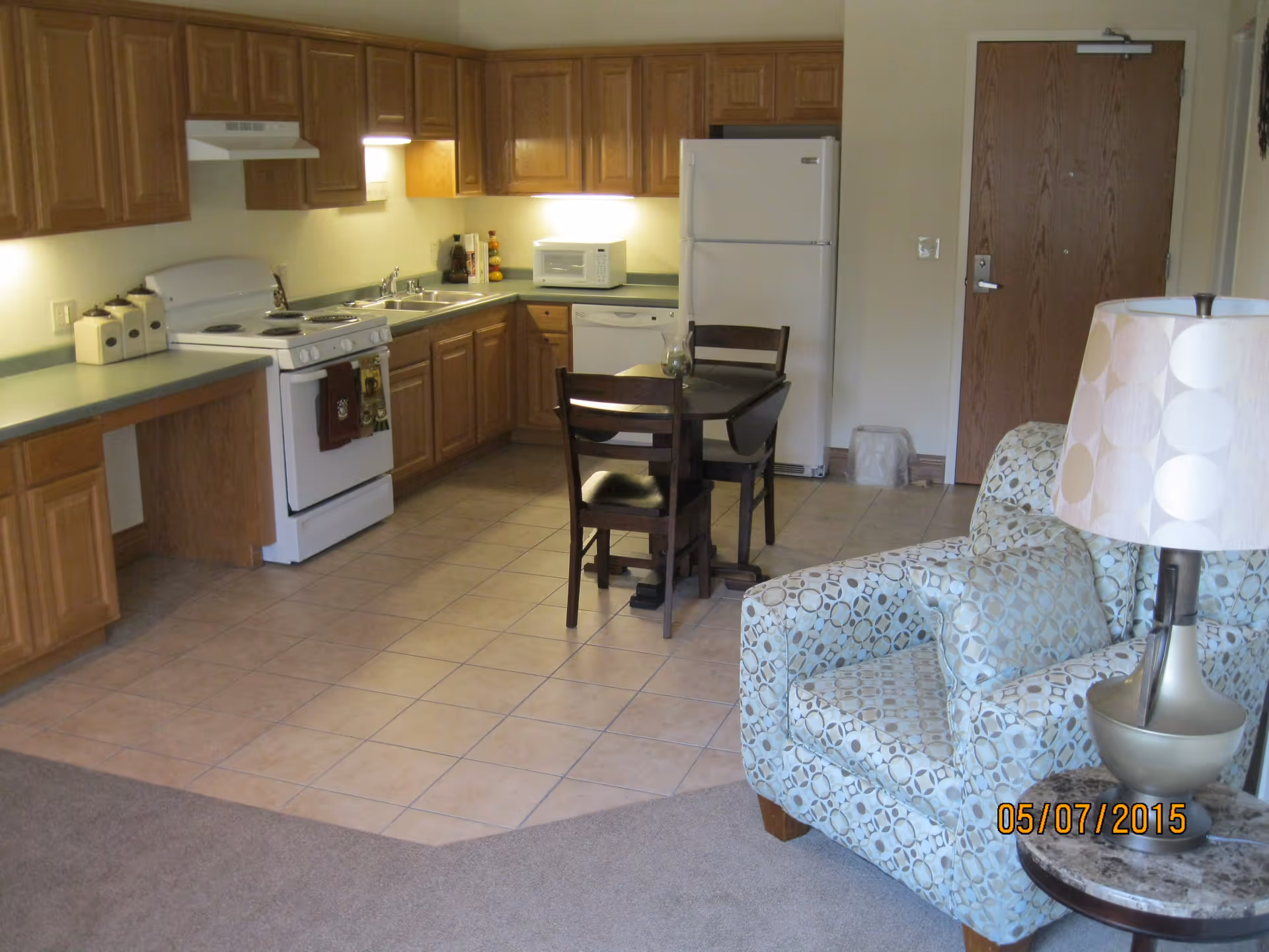 Interior view of a small apartment kitchen and living area with wooden cabinets, a white stove, refrigerator, microwave, and dishwasher. There is a small dining table with two chairs in the center. A patterned armchair and a lamp on a side table are visible in the foreground. The entrance door is in the background.