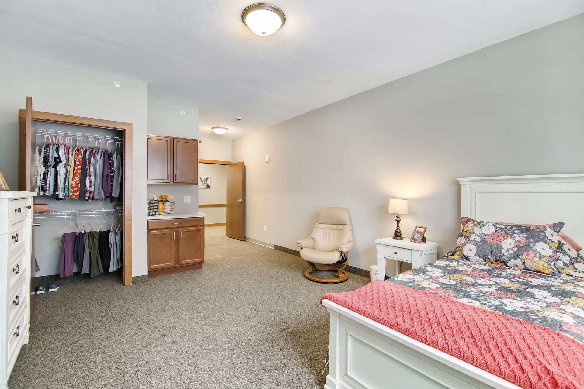 Bright bedroom with a floral bed and pink throw, nightstand and lamp, an armchair, open closet with hanging clothes, and a dresser.