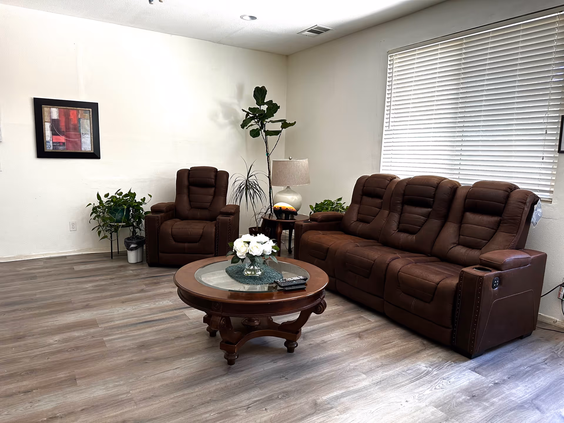 A bright living room with a brown three-seat recliner and matching armchair, a round wooden coffee table with flowers, houseplants, a lamp, and wall art.