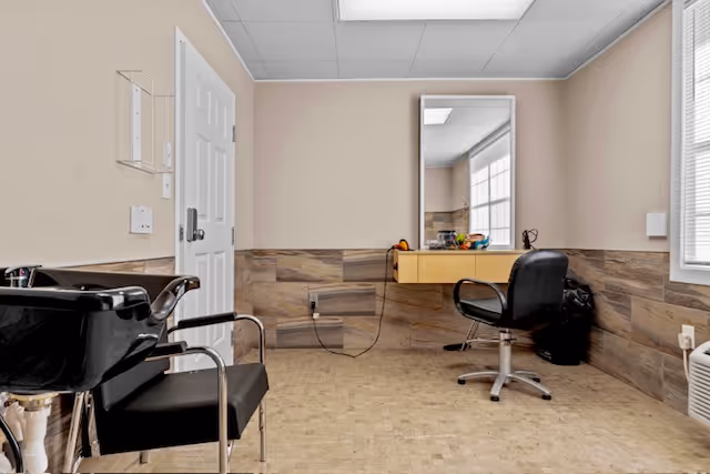 Interior view of a small hair salon area with a black shampoo chair and sink on the left, a styling chair in front of a wall-mounted mirror, and a window on the right letting in natural light. The walls have a beige upper half and a wood-textured lower half, with a tiled floor.