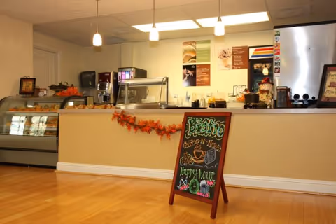 Bistro-style cafe counter with display cases, menu boards, hanging lights and a chalkboard sign in a senior living facility.