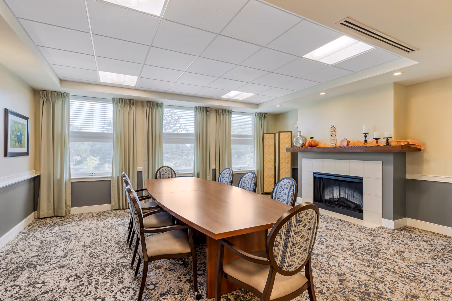 A well-lit dining room with a long wooden table surrounded by eight upholstered chairs. The room features large windows with beige curtains, a patterned carpet, and a fireplace decorated with candles and autumn-themed items. A framed picture hangs on one wall, and a folding screen is placed near the fireplace.
