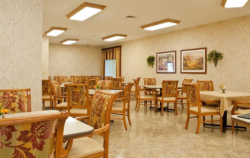 A dining room in Greenwood Meadows facility with multiple wooden tables and chairs featuring floral upholstery. The room has beige patterned wallpaper, framed landscape paintings on the wall, small flower arrangements on the tables, and ceiling lights providing bright illumination.