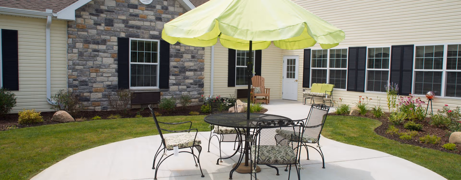 Outdoor patio area at Bickford Of Portage featuring a round metal table with four chairs, each with patterned cushions, and a large green umbrella providing shade. The patio is surrounded by a well-maintained lawn, flower beds, and the exterior walls of the building with windows and a door.