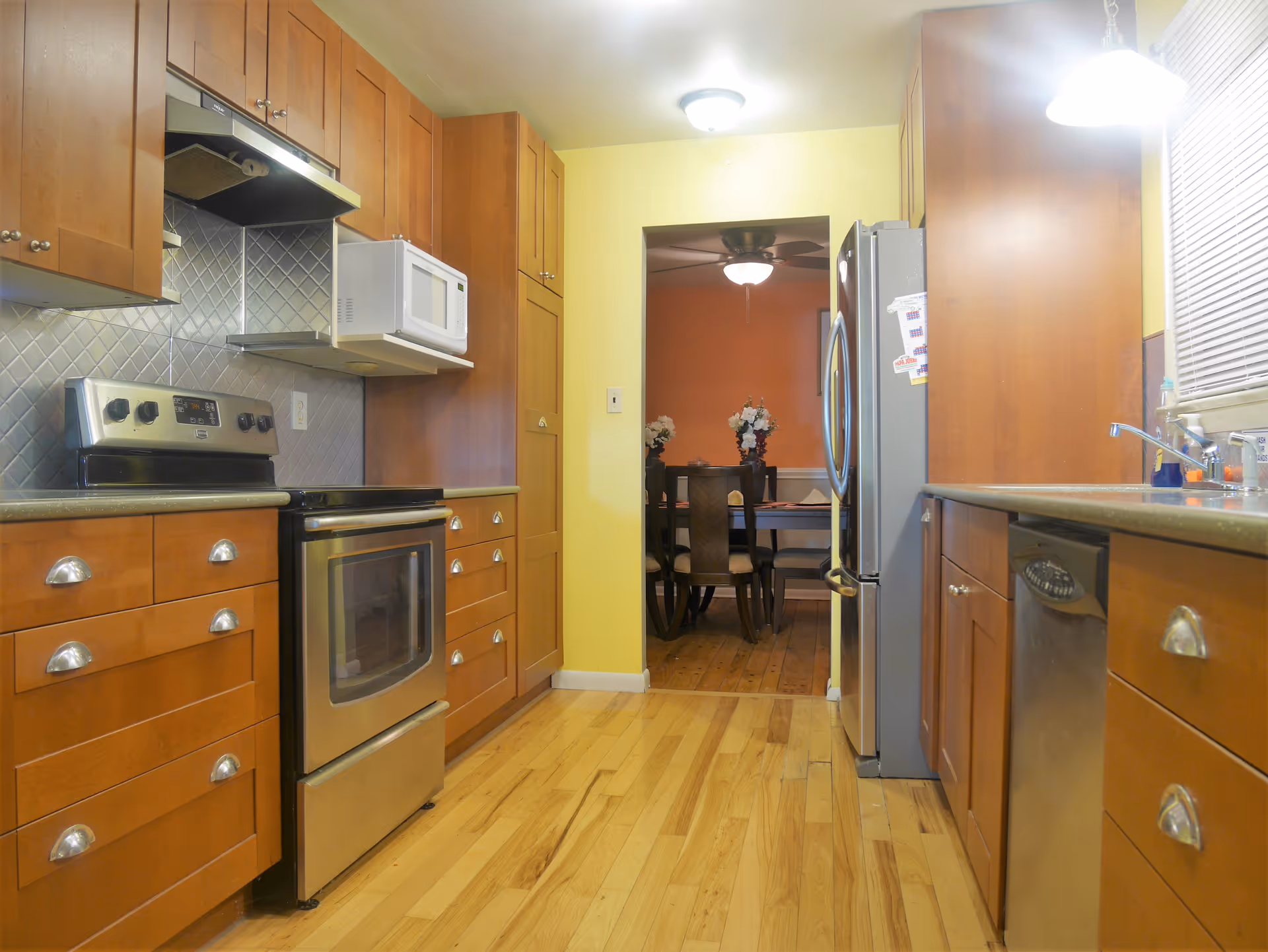 Bright galley kitchen with wooden cabinets, stainless steel appliances, and a view through a doorway to a dining room.