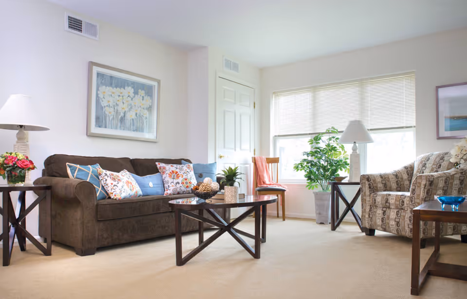 A bright living room with a brown sofa adorned with colorful pillows, a patterned armchair, two wooden side tables with lamps, a wooden coffee table with decorative items, a wooden chair with a coral throw, a large window with blinds, and a potted plant near the window.