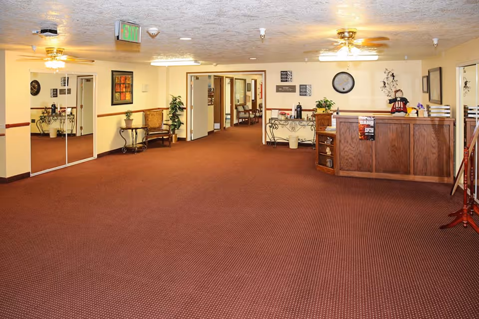 Interior view of a senior living facility lobby or common area with brown carpet, beige walls, ceiling fans, a wooden reception desk, decorative plants, chairs, wall art, and an exit sign above a doorway.