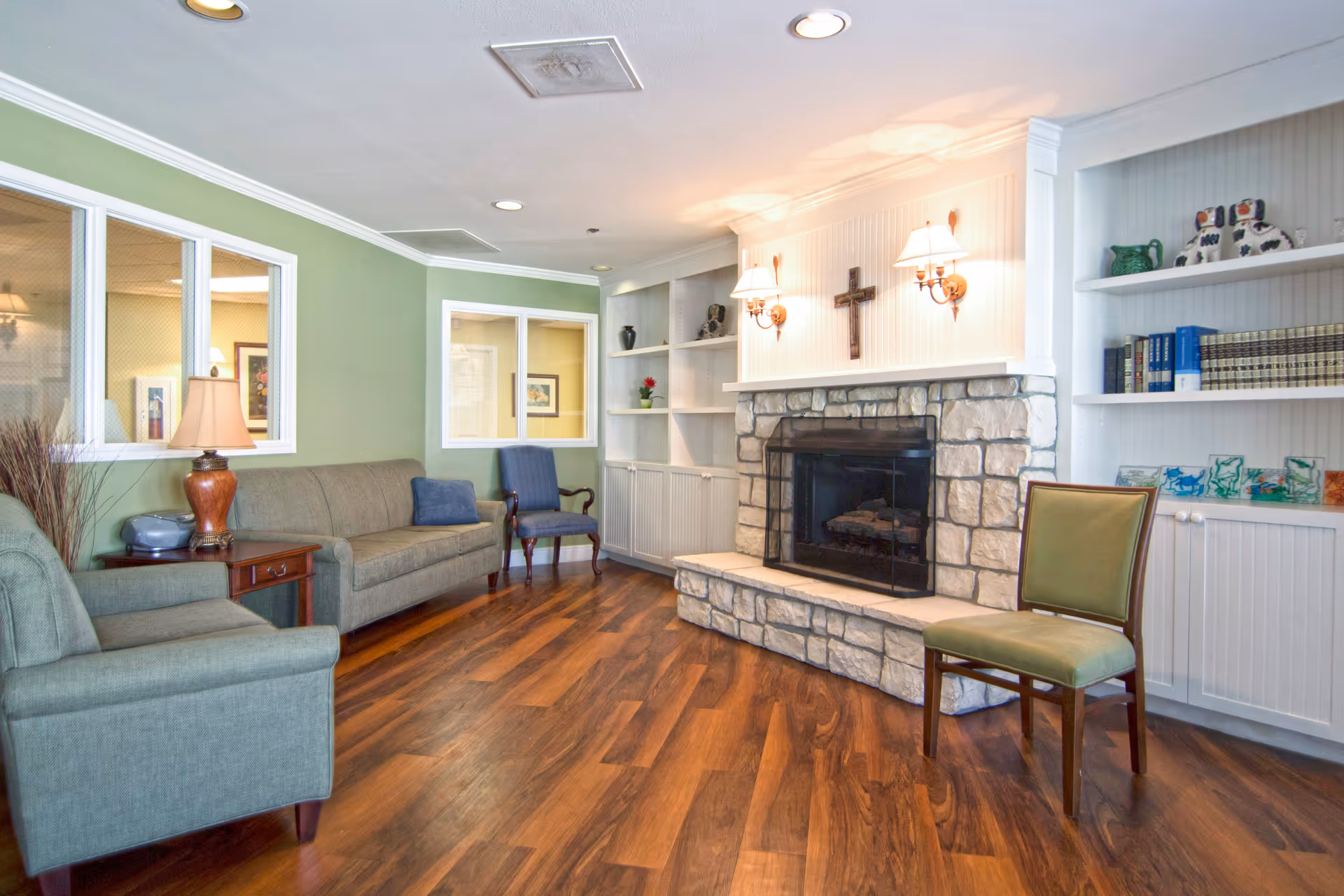 A cozy living room area with wooden flooring, a stone fireplace with a cross mounted above it, two wall lamps, built-in white shelves with books and decorative items, a green armchair, a green sofa, a wooden side table with a lamp, and a blue upholstered chair near a green wall with windows looking into another room.