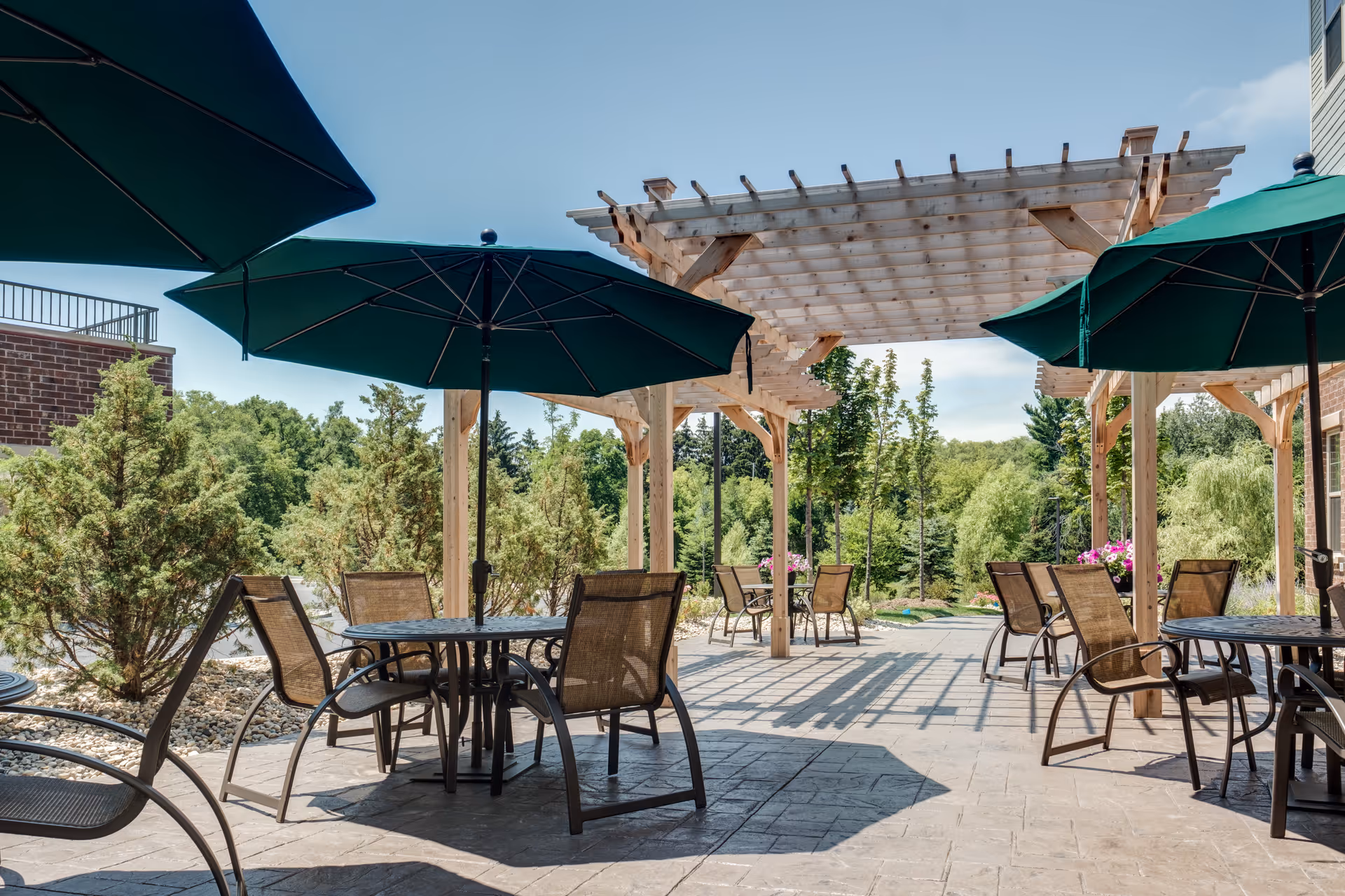 Outdoor patio with tables and chairs under green umbrellas and a wooden pergola overlooking trees and landscaping.