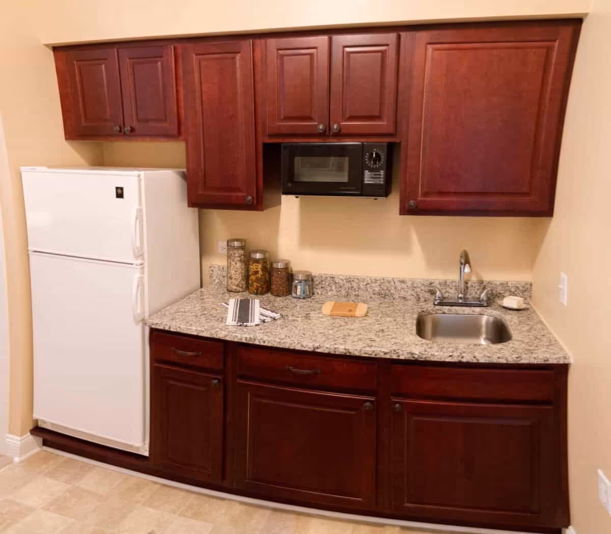 A small kitchen area with dark wooden cabinets, a white refrigerator, a black microwave mounted above the granite countertop, a stainless steel sink, and several glass jars containing dry food items on the counter.