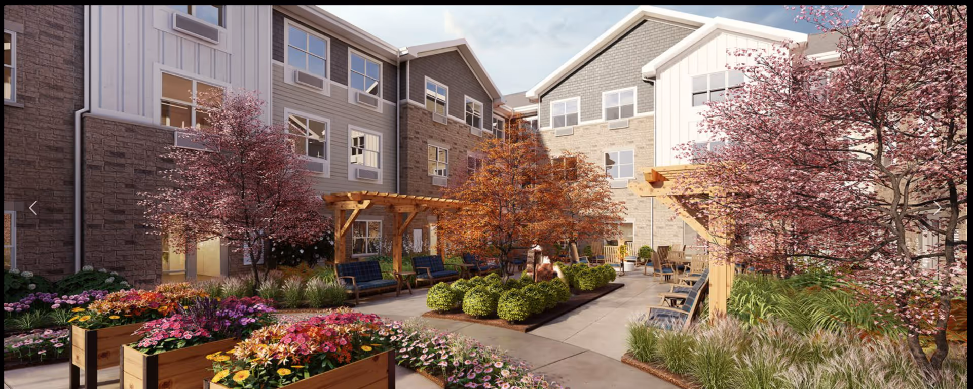 Sunny landscaped courtyard with raised flower beds, seating areas, pergolas and flowering trees beside a three-story senior living building.
