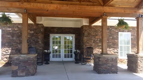 Covered entrance area of a building with stone pillars and wooden beams. There are two black rocking chairs on either side of a set of double glass doors. Hanging plants are suspended from the wooden ceiling.