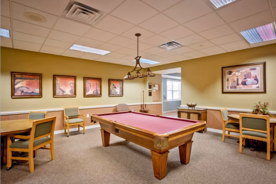 Recreation room with a pool table, surrounding chairs and tables, framed artwork, and overhead lighting.