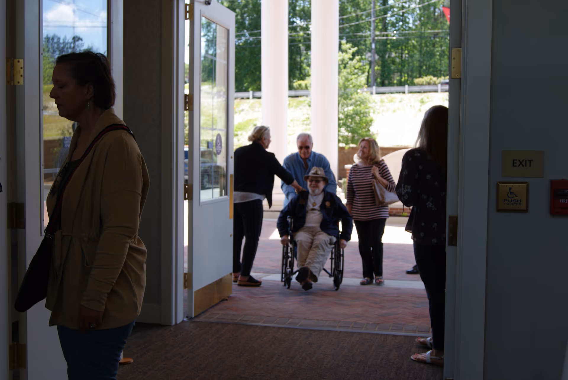 View from inside a building looking out through open double doors to a group of people outside. One person is in a wheelchair being pushed by another, while three other people stand nearby on a brick patio with greenery and trees in the background.