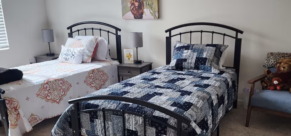 Two twin beds with patterned quilts and metal headboards, a nightstand with lamps between them, and a chair with stuffed animals in a bright bedroom.