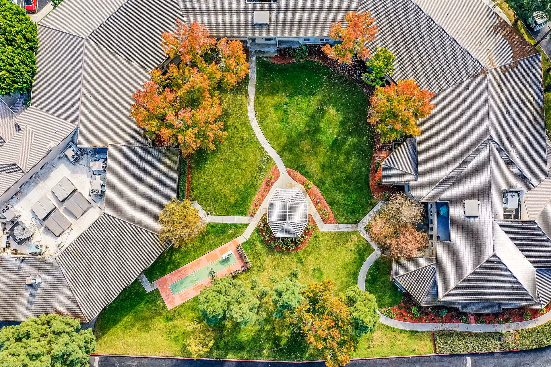 Aerial view of Arcadia Place Senior Living showing a large green courtyard with a central gazebo surrounded by pathways and trees with autumn foliage. The courtyard is enclosed by buildings with gray roofs and there is a shuffleboard court on the left side.