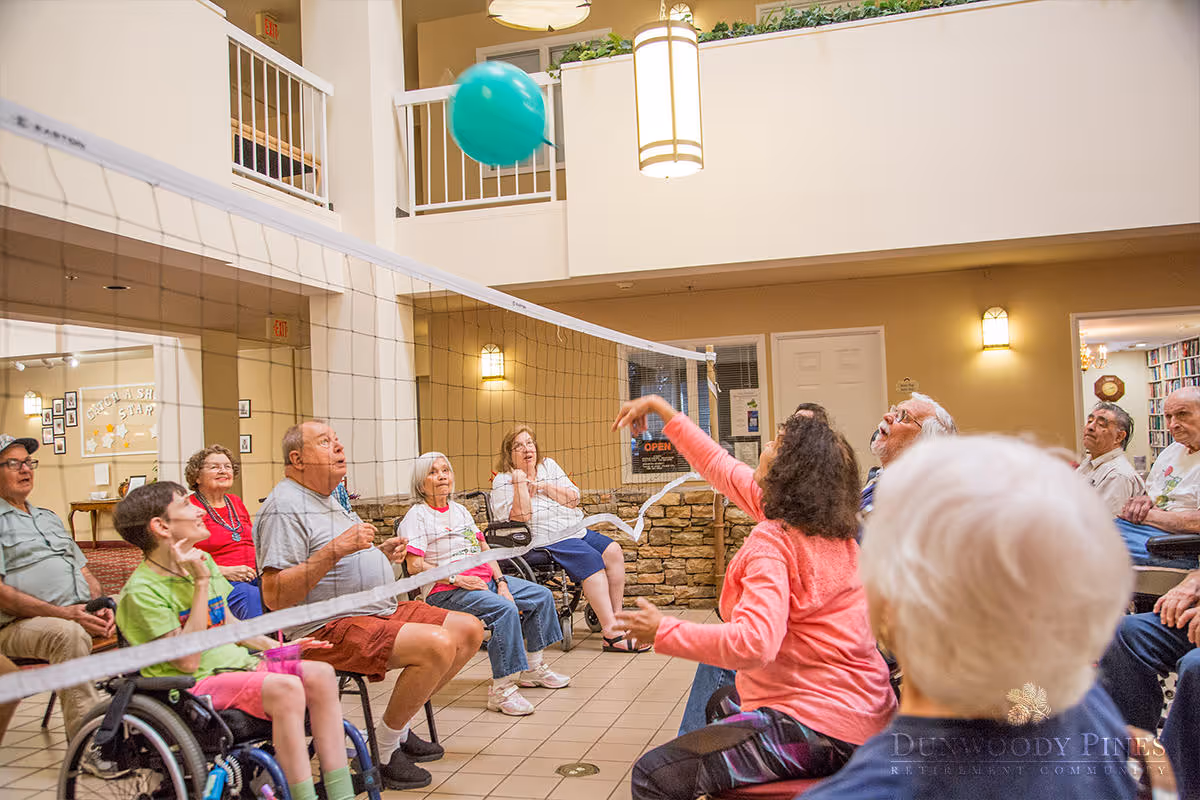 A group of elderly people and a child in a wheelchair playing seated volleyball indoors in a common area with a volleyball net and a green ball in mid-air. The room has a high ceiling with hanging lights and a second-floor balcony overlooking the area.
