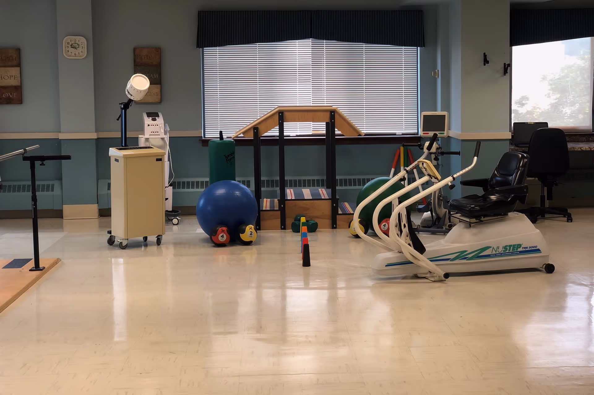 A physical therapy or exercise room with various equipment including a NuStep recumbent cross trainer, exercise balls, dumbbells, parallel bars, and a step platform. The room has light blue walls, a large window with blinds, and a clock on the wall.