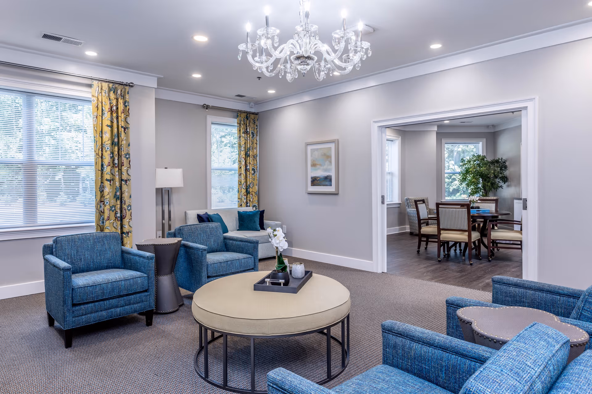 A bright and spacious senior living facility common area featuring blue upholstered armchairs arranged around a large round beige ottoman with a decorative tray and orchid. The room has large windows with yellow floral curtains, a white sofa with blue cushions, a floor lamp, and a crystal chandelier overhead. An adjacent room with a wooden dining table and chairs is visible through a wide doorway, with a large green plant near the window.