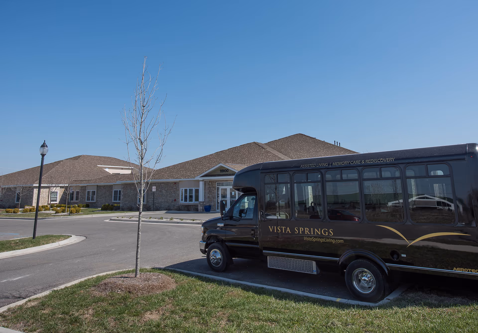 Exterior view of Vista Springs Washington Place facility with a black shuttle bus parked in front. The building has a stone facade and a brown roof under a clear blue sky. A small tree and a street lamp are visible near the parking lot.