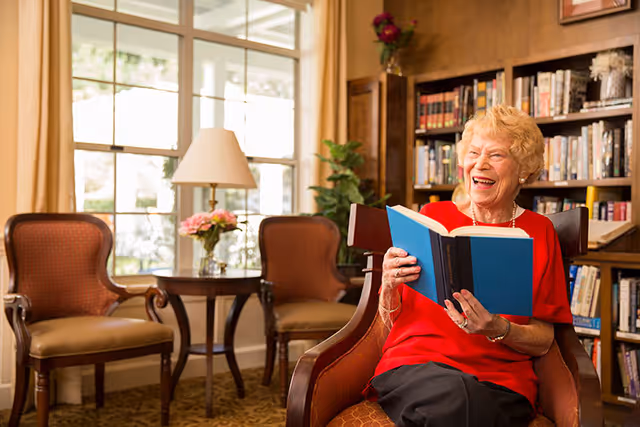 An elderly woman wearing a red top is sitting in a comfortable chair in a cozy room with bookshelves and large windows. She is smiling and holding an open book. The room has warm lighting, two empty armchairs, a small table with a lamp and flowers, and a plant near the window.