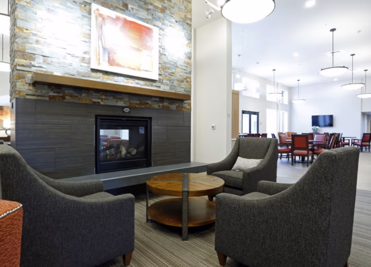 A cozy seating area with three dark gray armchairs arranged around a round wooden coffee table in front of a modern stone fireplace. In the background, there is a dining area with multiple tables and red chairs, bright white walls, and large windows letting in natural light.