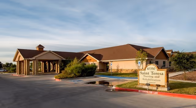 Exterior view of Saint Teresa Nursing and Rehabilitation Center building with a covered entrance, parking spaces, and a sign displaying the facility's name and address.