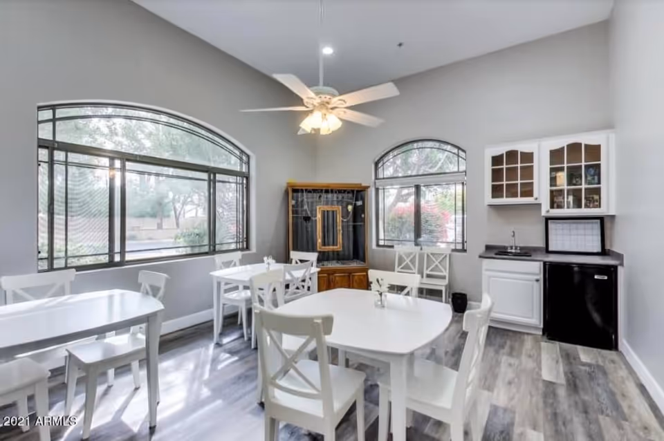 A bright dining area with white tables and chairs, large arched windows letting in natural light, a ceiling fan with lights, and a small kitchenette with white cabinets, a sink, and a mini refrigerator.