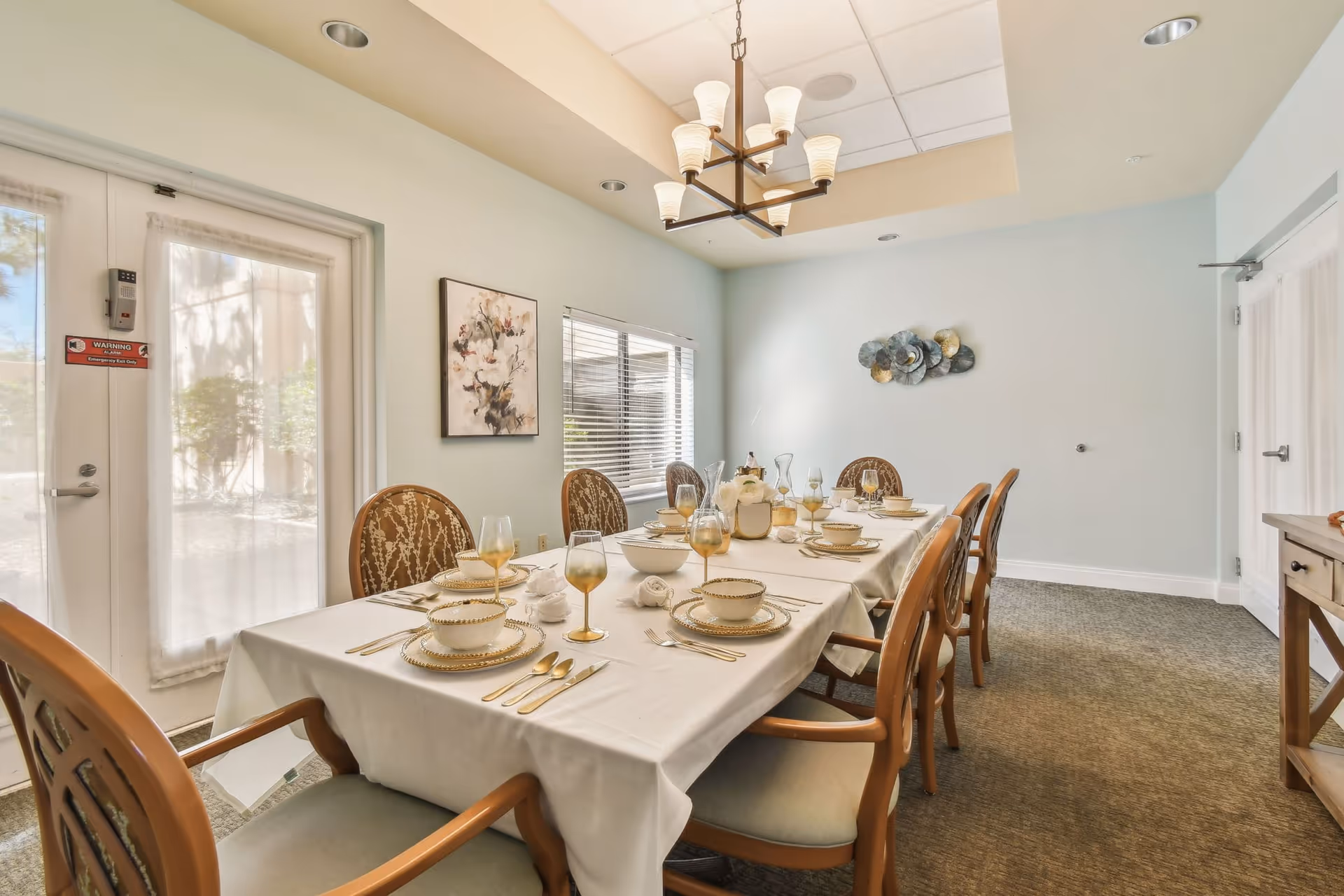 A dining room with a long table set for eight people, featuring elegant tableware including plates, bowls, glasses, and cutlery. The room has light blue walls, a window with blinds, a decorative wall art piece, and a chandelier hanging from the ceiling. There is a door with sheer curtains on the left side and a wooden sideboard on the right.