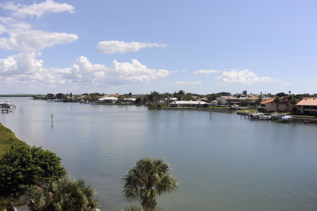 A calm waterway with houses and docks along the shoreline under a blue sky with scattered clouds. Palm trees and other greenery are visible near the water.