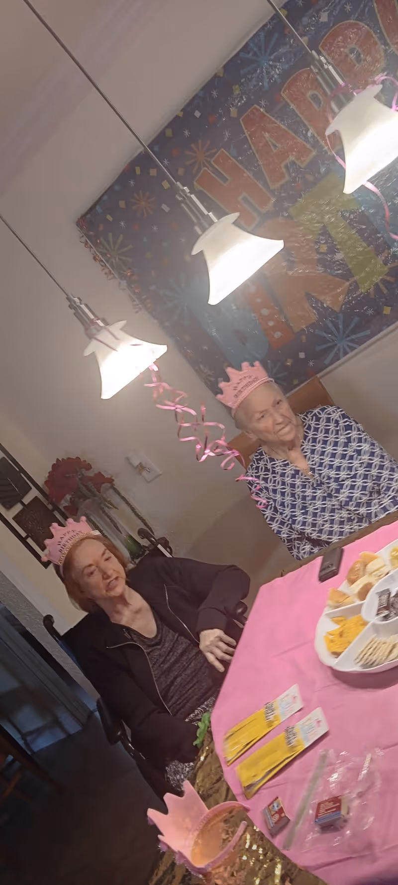 Two elderly women wearing pink birthday crowns sitting at a table covered with a pink tablecloth. The table has snacks and party items on it. Behind them is a colorful 'Happy Birthday' banner and two hanging pendant lights.