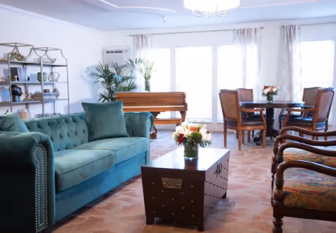 A bright living room with a green velvet sofa, a wooden coffee table with a flower vase, a round dining table with four chairs, a piano near large windows with sheer curtains, and a bookshelf with decorative items and plants.