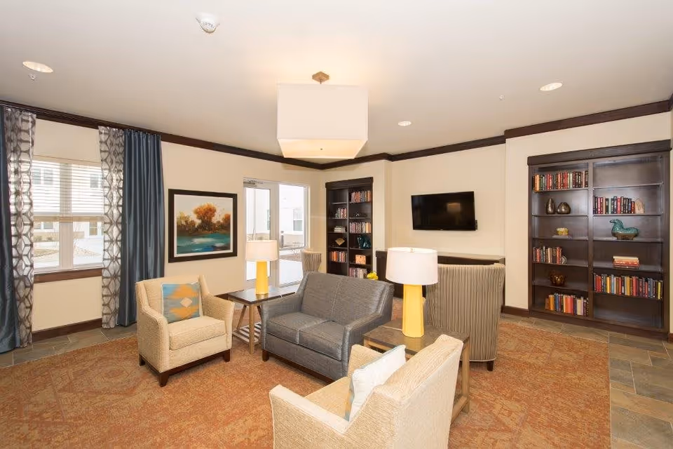 A cozy living room area with a gray loveseat, two beige armchairs, and a striped armchair arranged around two side tables with yellow lamps. The room features two large dark wood bookshelves filled with books and decorative items, a wall-mounted flat screen TV, a large window with patterned curtains, and a framed landscape painting on the wall. The floor is covered with a patterned rug and the walls have dark wood trim.
