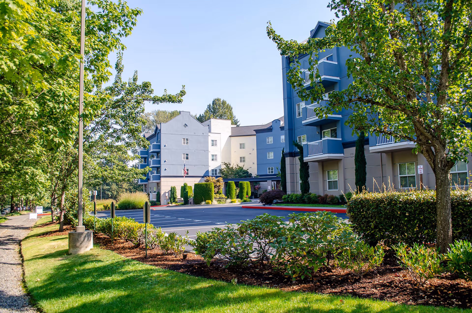 Exterior view of Chateau Valley Center Retirement facility showing a multi-story building with blue and beige walls, surrounded by green trees, shrubs, and a well-maintained lawn under a clear blue sky. There is an empty parking lot in front of the building.