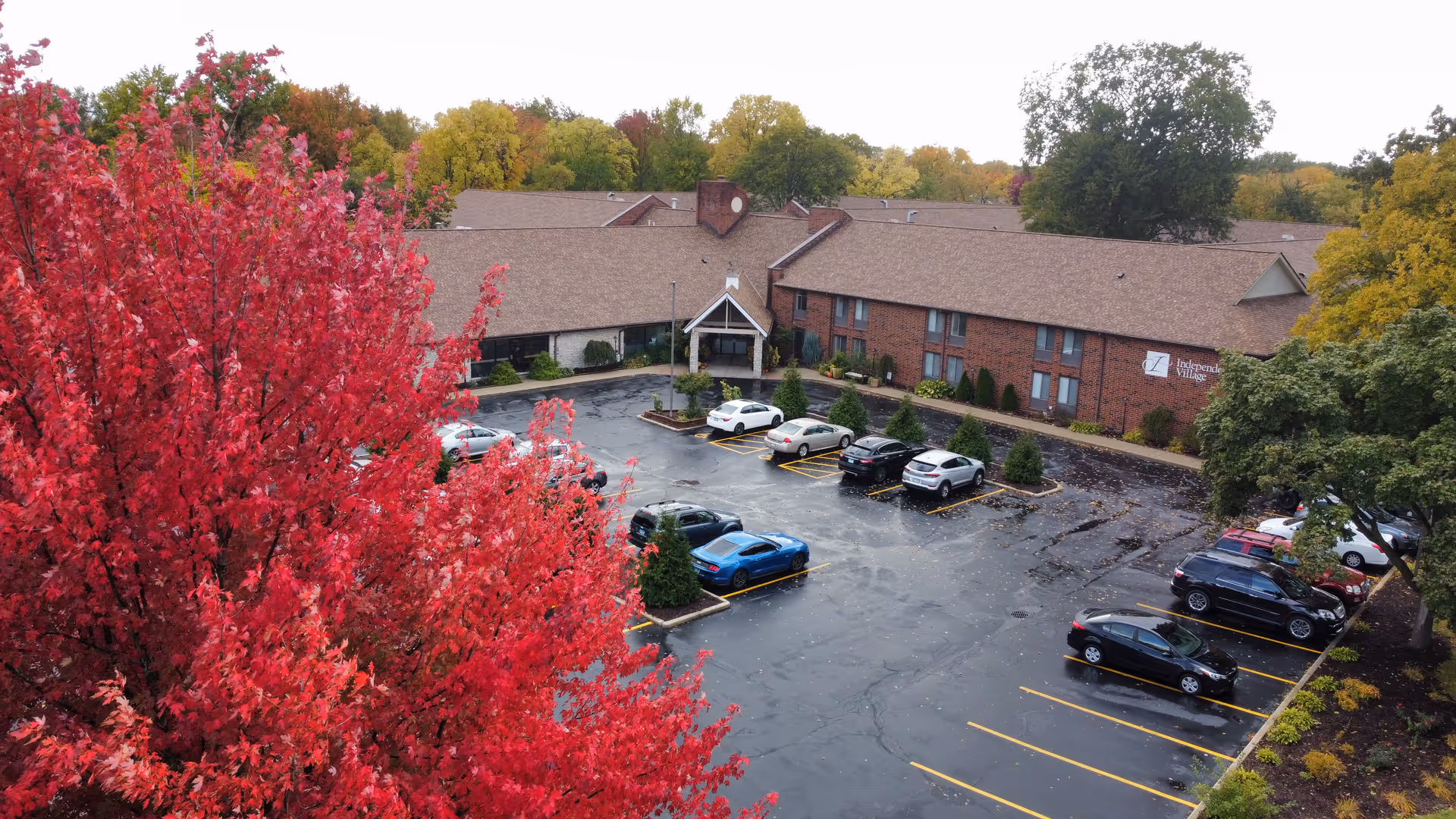 Aerial view of Independence Village senior living facility showing a large parking lot with several parked cars, surrounded by trees with autumn foliage including vibrant red and green leaves. The building is a two-story brick structure with a peaked entrance and a sign that reads Independence Village.