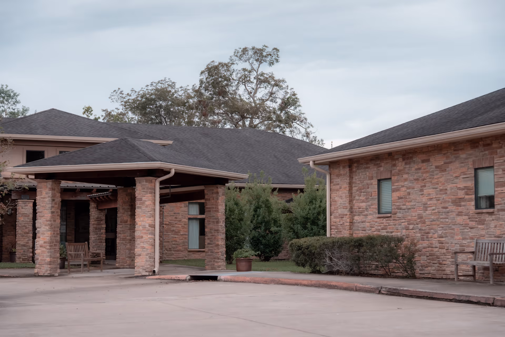 Exterior view of a senior living facility with brick buildings, a covered entrance supported by brick columns, benches, shrubs, and trees under a cloudy sky.