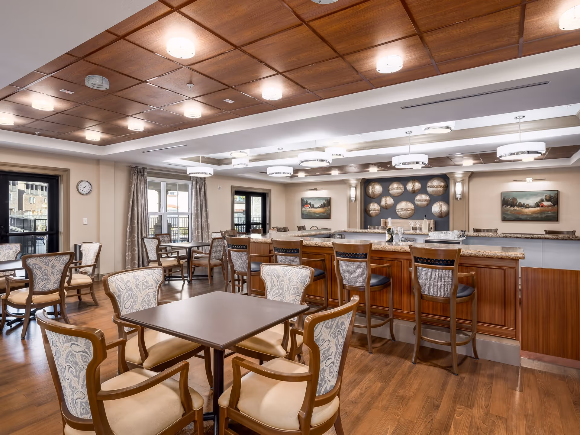 A spacious dining area with multiple tables and chairs, a wooden floor, and a ceiling with recessed lighting and wooden panels. There is a large counter with bar stools, decorative wall art, and windows with curtains allowing natural light to enter.
