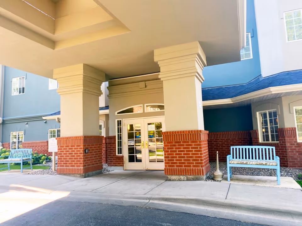 Entrance of a senior living facility with double glass doors framed by large beige columns with red brick bases. Two light blue benches with cushions are placed on either side of the entrance. The building exterior is painted in shades of blue and beige with multiple windows visible.