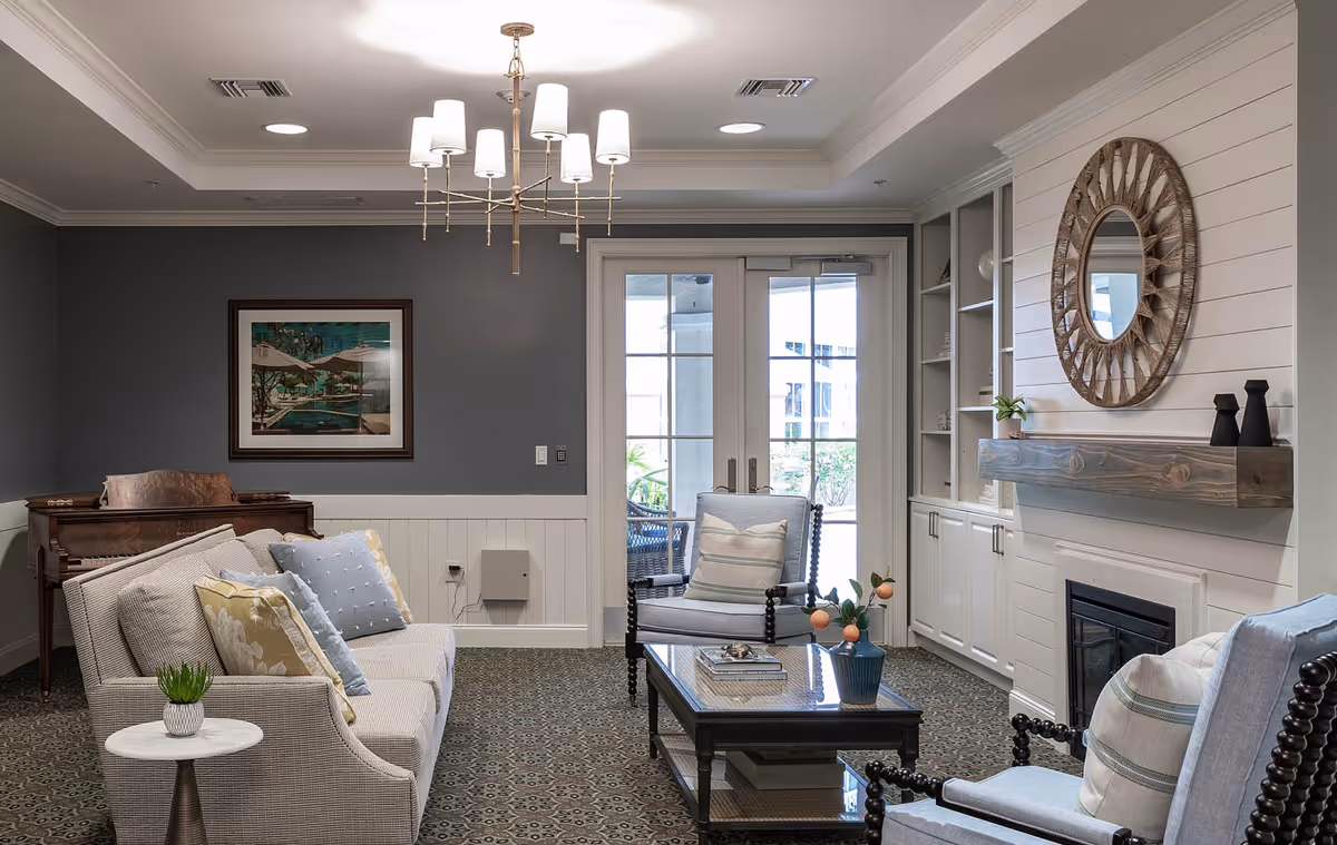 Well-appointed living room with sofas and armchairs around a coffee table, a fireplace with a round mirror above it, a piano in the corner, and French doors leading outside.