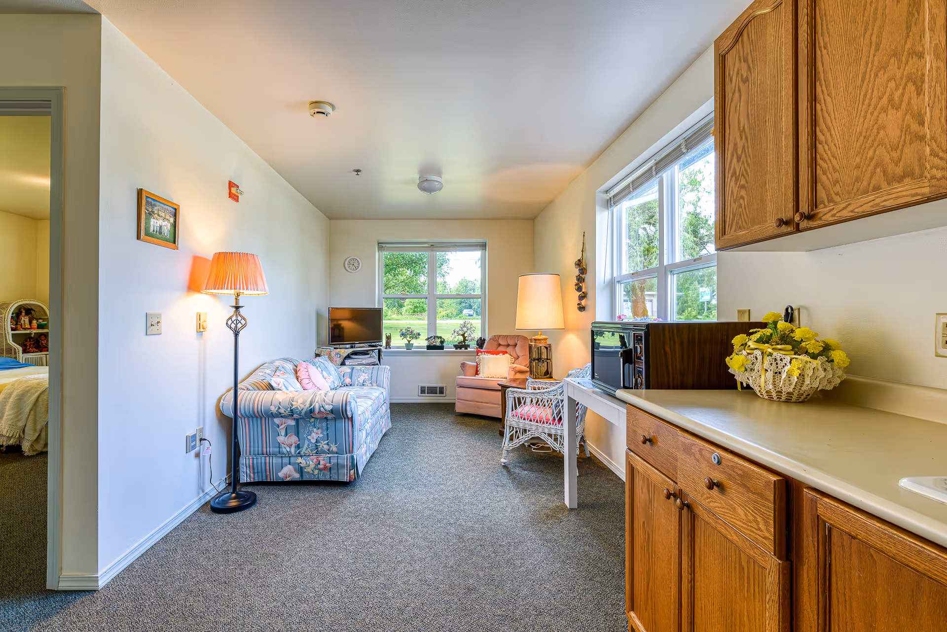 Cozy assisted-living studio living area with a floral sofa, armchair, TV, lamps, and a small kitchenette with wooden cabinets by a large window.