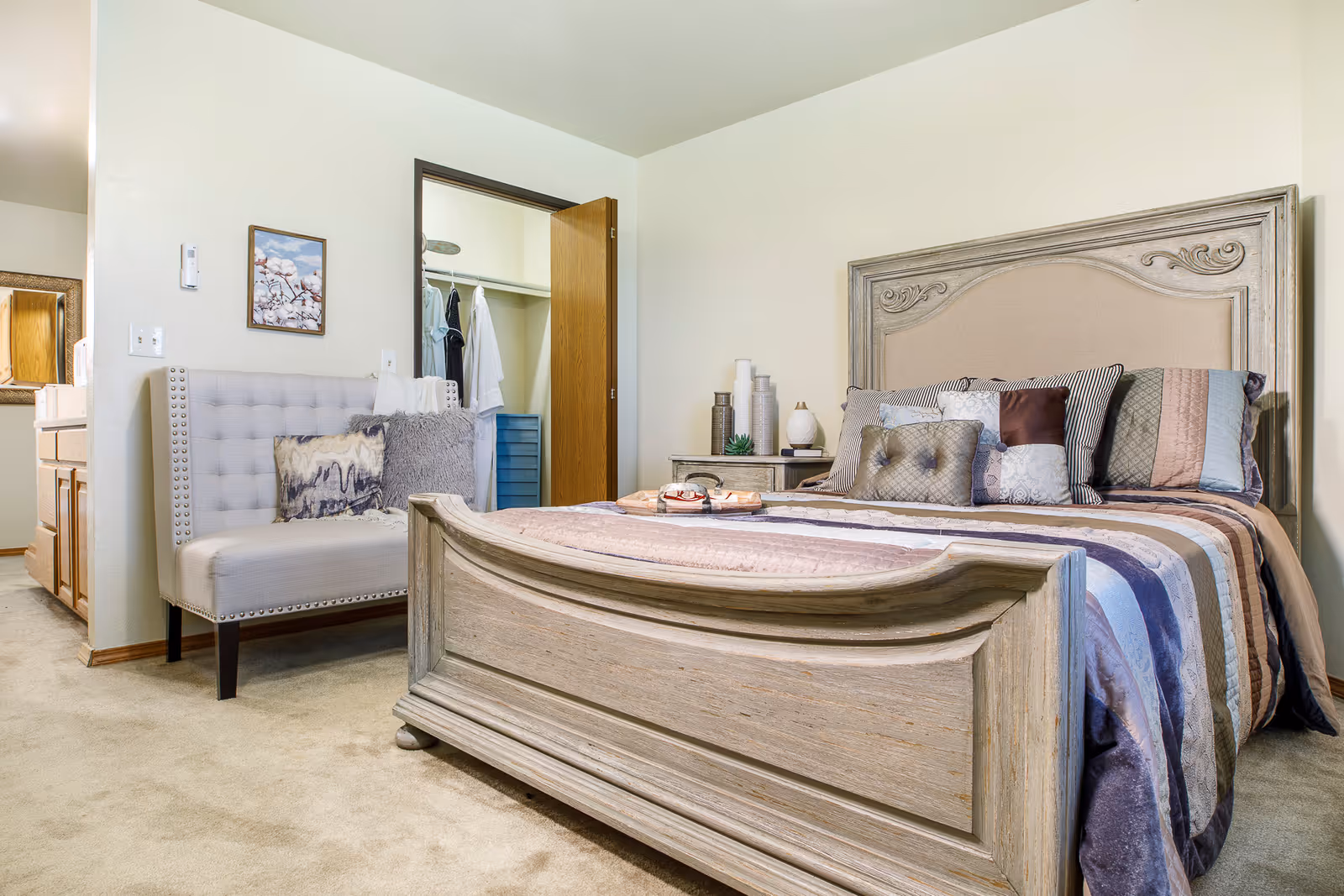 A bedroom in a senior living facility featuring a large wooden bed with a decorative headboard and multiple pillows. Next to the bed is a nightstand with decorative items. There is a tufted bench with pillows against the wall, and an open door reveals a closet with hanging clothes. The room has light-colored walls and carpeted flooring.