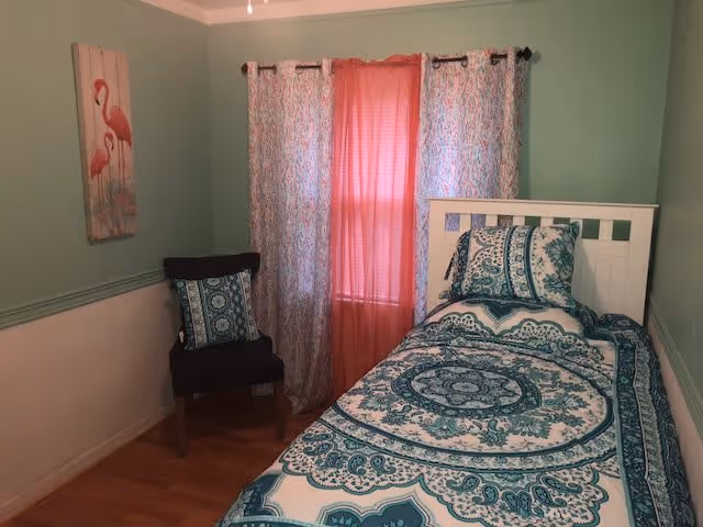 A small bedroom with a single bed featuring a blue and white patterned bedspread and matching pillow. Next to the bed is a dark chair with a decorative cushion. The walls are painted light green with a white lower half, and there is a window with pink and patterned curtains. A flamingo-themed wall art hangs on the left wall.