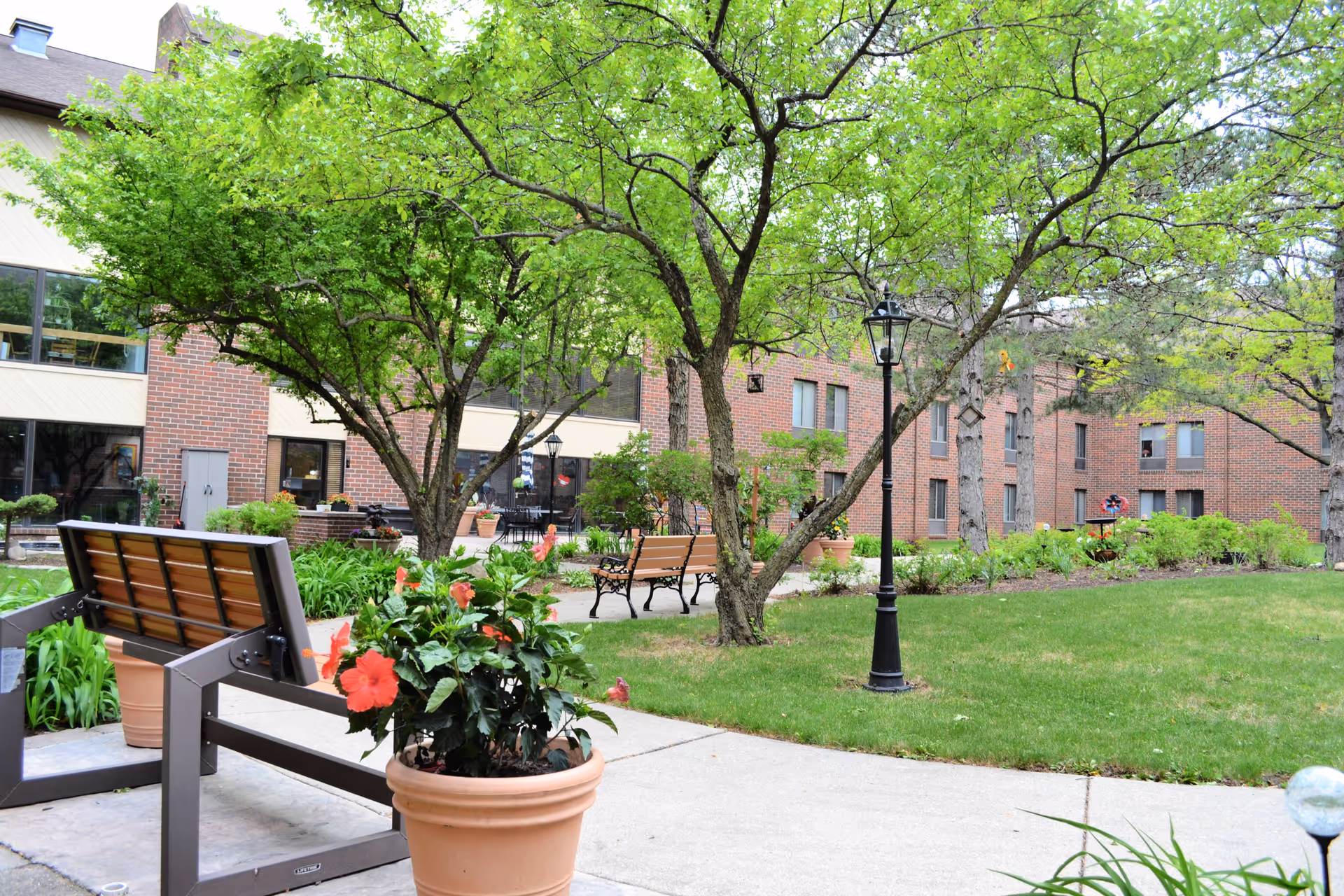 Courtyard with benches, potted flowers, lamp posts and trees in front of a brick residential building.