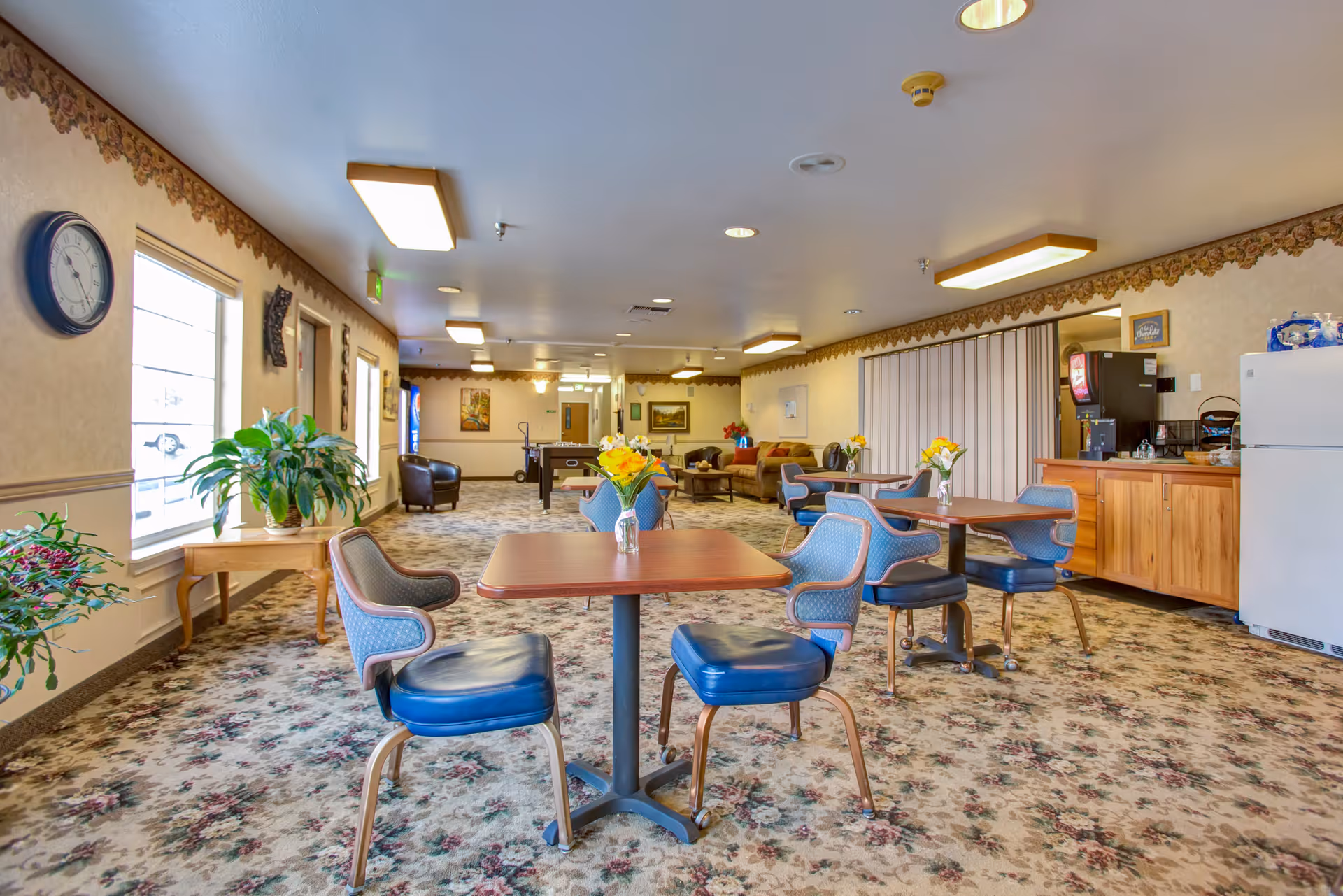 A senior living facility common area with several tables and blue cushioned chairs arranged around them. Each table has a small vase with yellow flowers. The room has floral patterned carpet, beige walls with a decorative border near the ceiling, and multiple ceiling lights. There are large windows on the left side letting in natural light, a clock on the wall, some potted plants, and a kitchenette area with a refrigerator and coffee machine on the right. In the background, there are more seating areas and a foosball table.