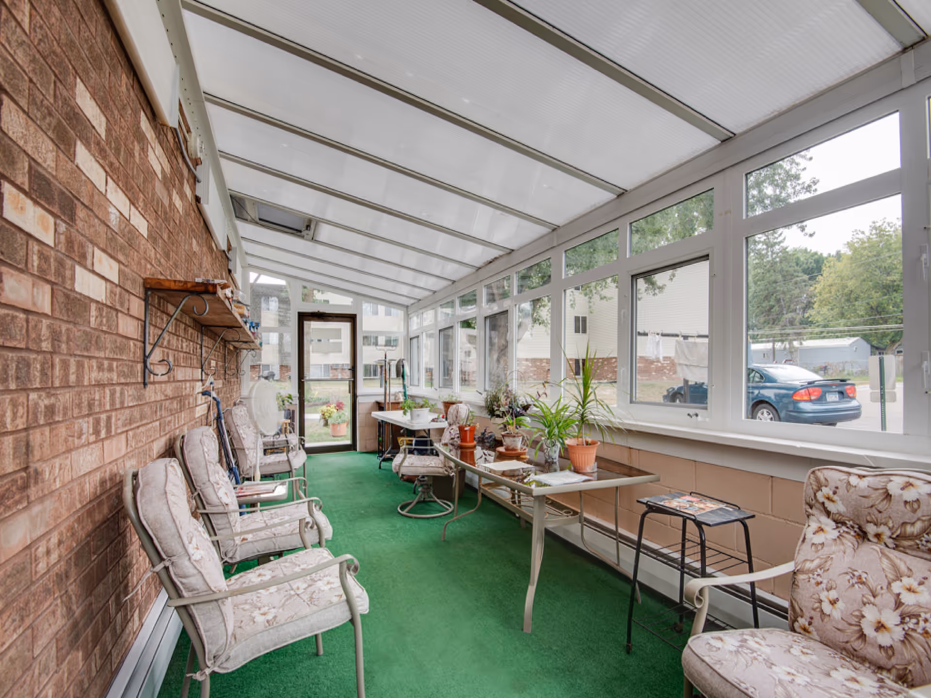 A sunroom with a green carpet floor, several cushioned chairs with floral patterns, a glass-top table with potted plants, and large windows letting in natural light. The room has a brick wall on one side and a white framed glass door at the end leading outside.