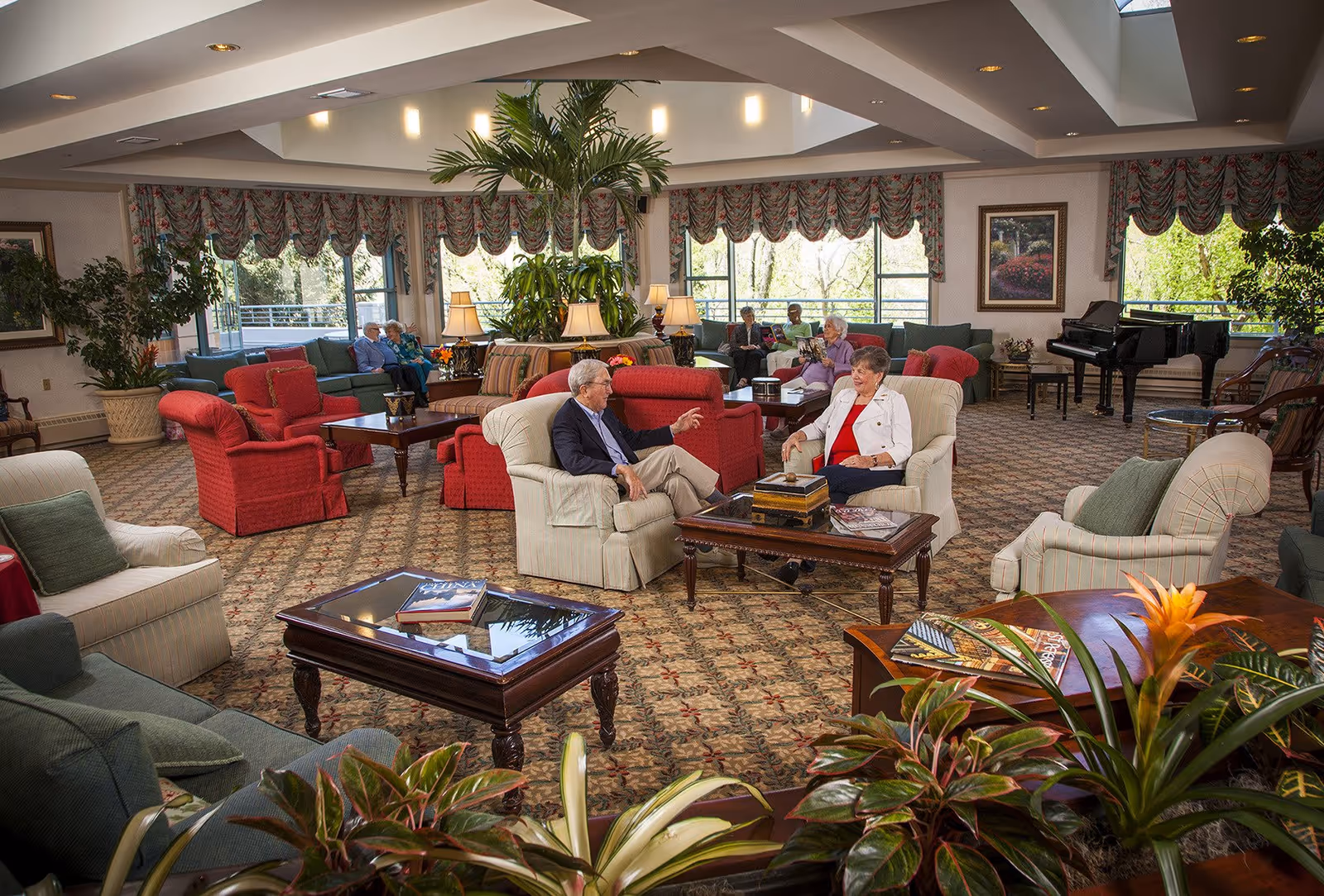 A spacious and well-lit senior living facility common area with comfortable armchairs and sofas arranged around coffee tables. Several elderly people are sitting and conversing or reading. The room features large windows with floral valances, indoor plants, table lamps, patterned carpet, and a grand piano in the background.