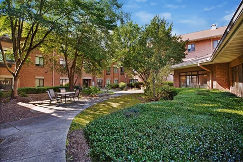 Sunlit landscaped courtyard with a curved paved walkway, patio table and chairs, trees and shrubs in front of brick residential buildings.